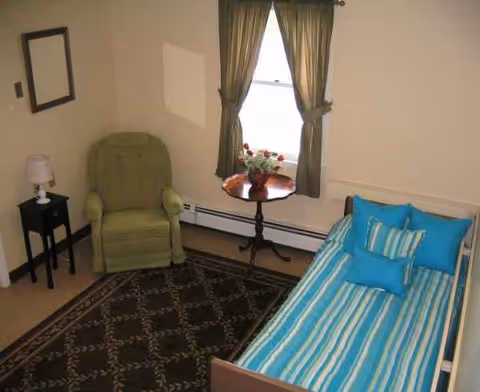 Cozy assisted-living bedroom with striped bed, green chair, window, and small table with flowers.