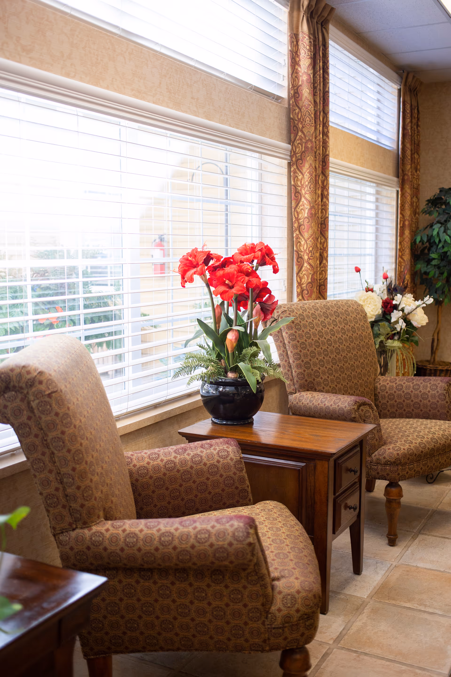 A cozy sitting area with two patterned armchairs and a wooden side table between them. On the table is a black vase with bright red flowers. Large windows with white blinds and patterned curtains let in natural light. There is also a plant with white flowers in the background.