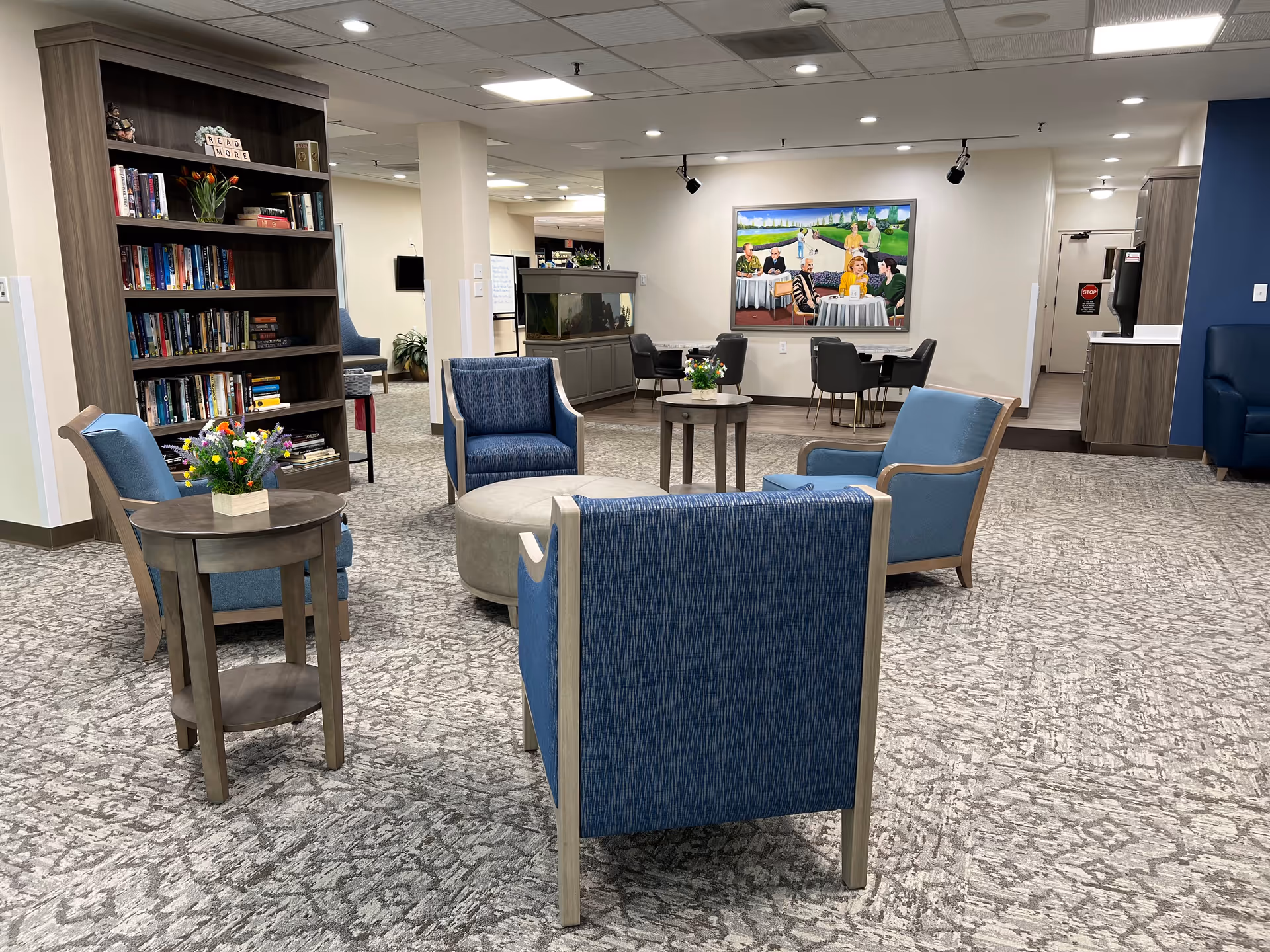 A cozy lobby area with blue chairs, a small table with flowers, a bookshelf filled with books, and a mural on the wall depicting a social gathering.