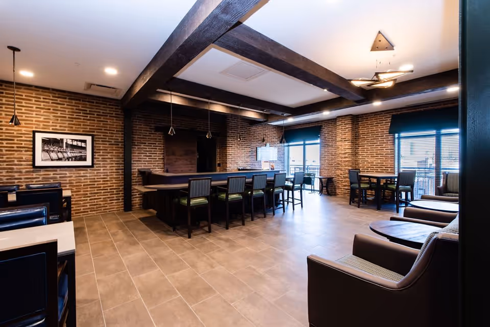 Spacious brick-walled lounge and dining area with a central bar, multiple tables and chairs, and large windows.