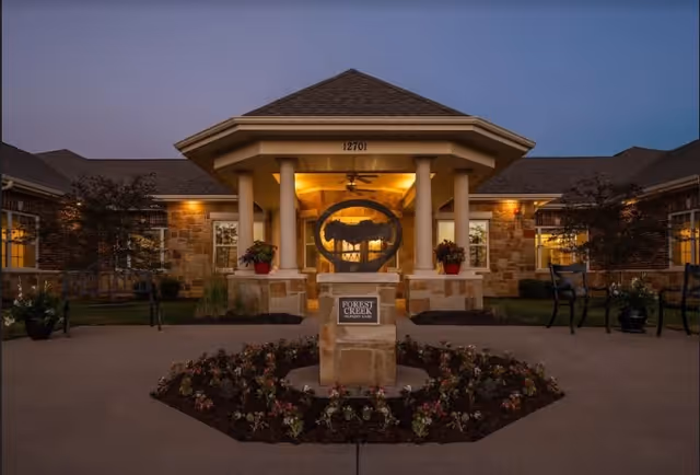 Exterior view of a senior living facility entrance at dusk with warm lighting. The building features stone walls, a covered entryway supported by columns, and a landscaped flower bed in front. A sign in the flower bed reads 'Forest Creek Memory Care'.