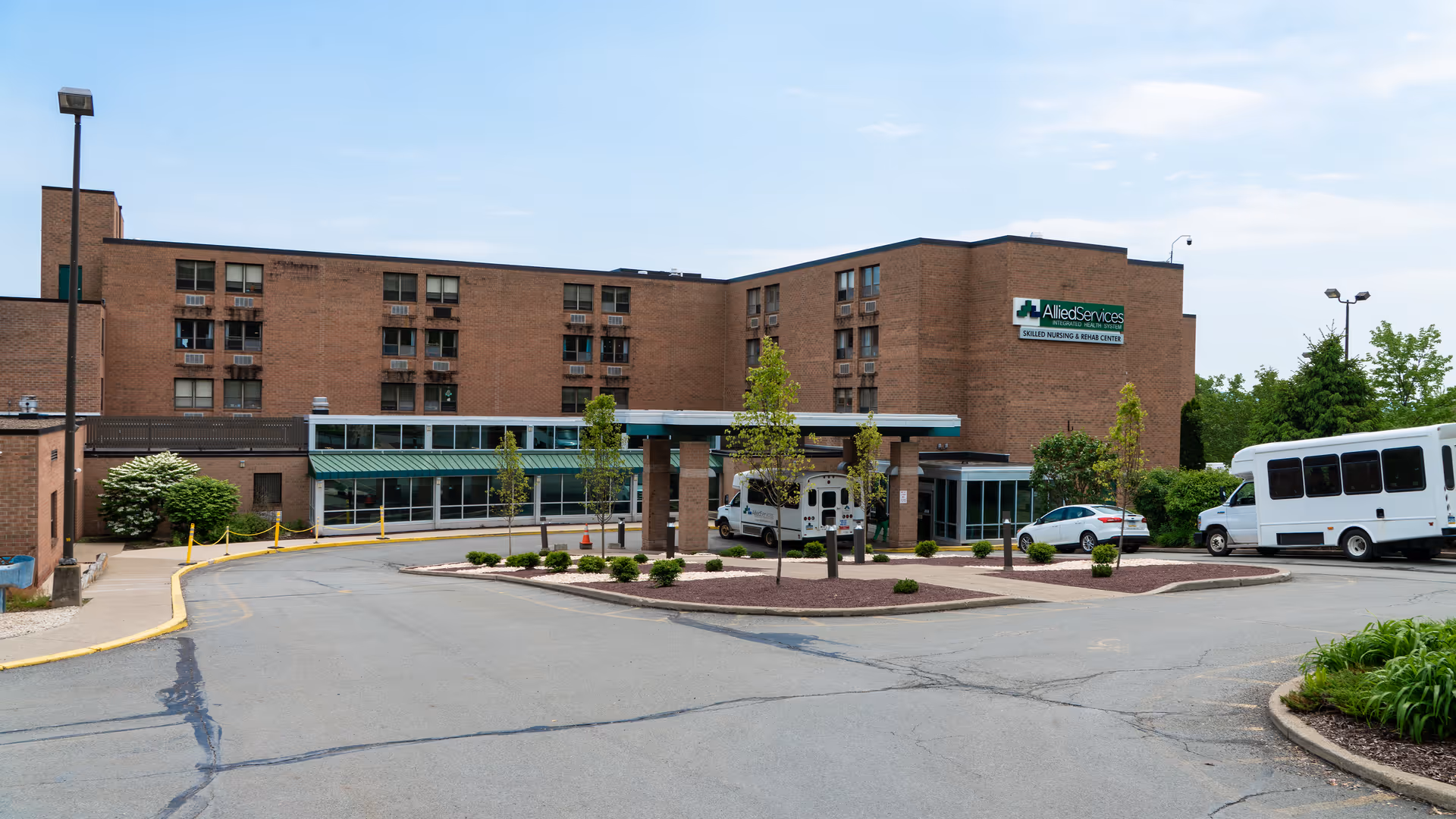 Exterior view of Allied Services Skilled Nursing & Rehabilitation Center, a multi-story brick building with a covered entrance driveway. Several vehicles, including a white van and a white bus, are parked near the entrance. The area is landscaped with small trees and shrubs, and the sky is clear with some clouds.