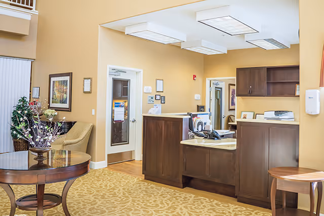 Interior view of a senior living facility reception area with a wooden front desk, beige walls, framed artwork, a round wooden table with a floral arrangement, and comfortable seating. The area is well-lit with ceiling lights and has a patterned carpet.
