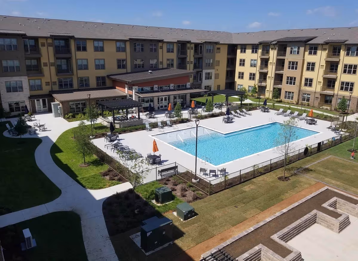 Aerial view of a senior living facility courtyard featuring a large rectangular swimming pool surrounded by lounge chairs and tables with umbrellas. The courtyard is enclosed by a multi-story building with numerous windows and balconies. There are paved walkways, green lawns, and small trees around the pool area under a clear blue sky.