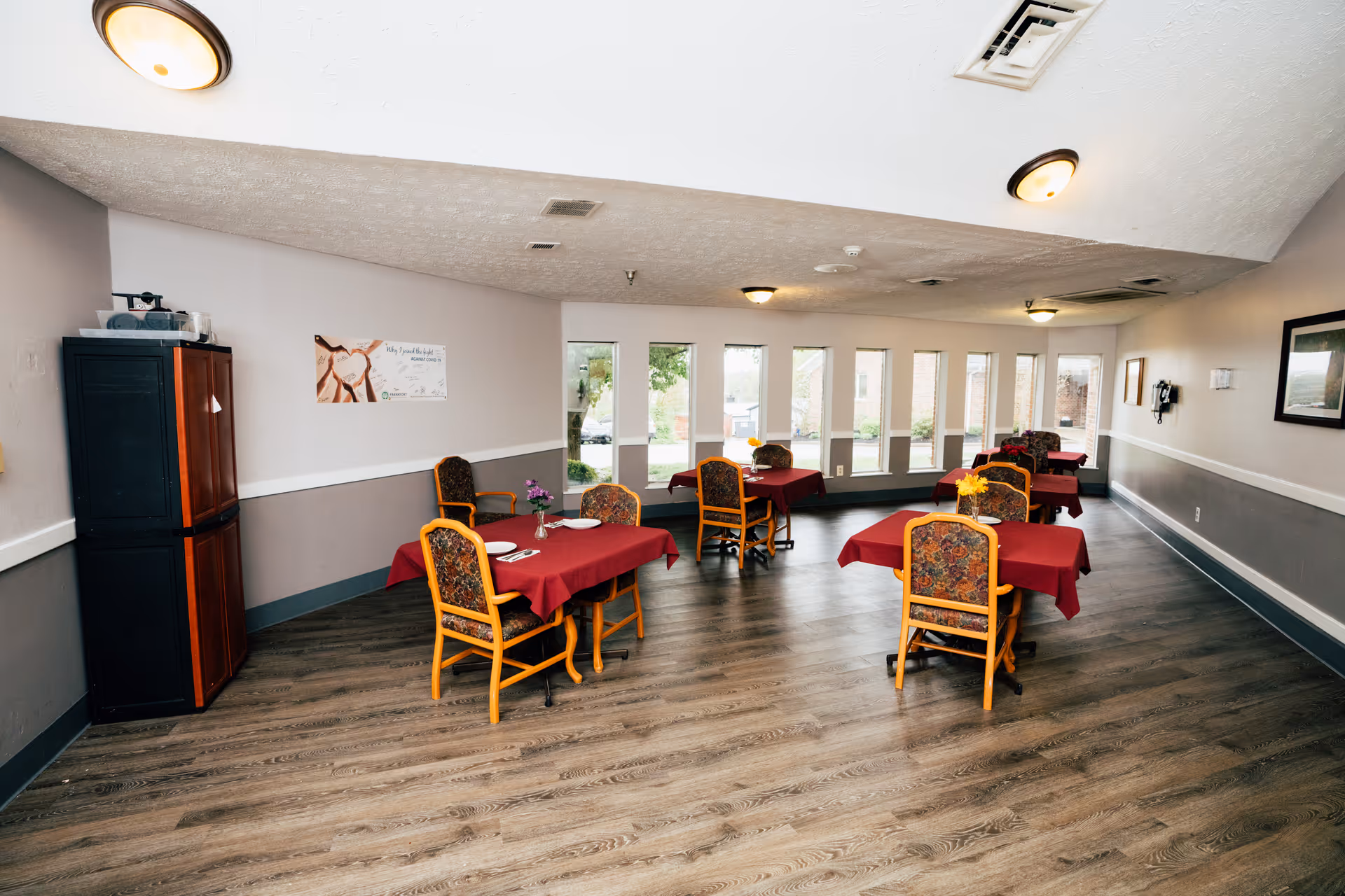 Spacious communal dining room with several tables covered in red tablecloths and patterned chairs, wood-look flooring and a row of windows along one wall.