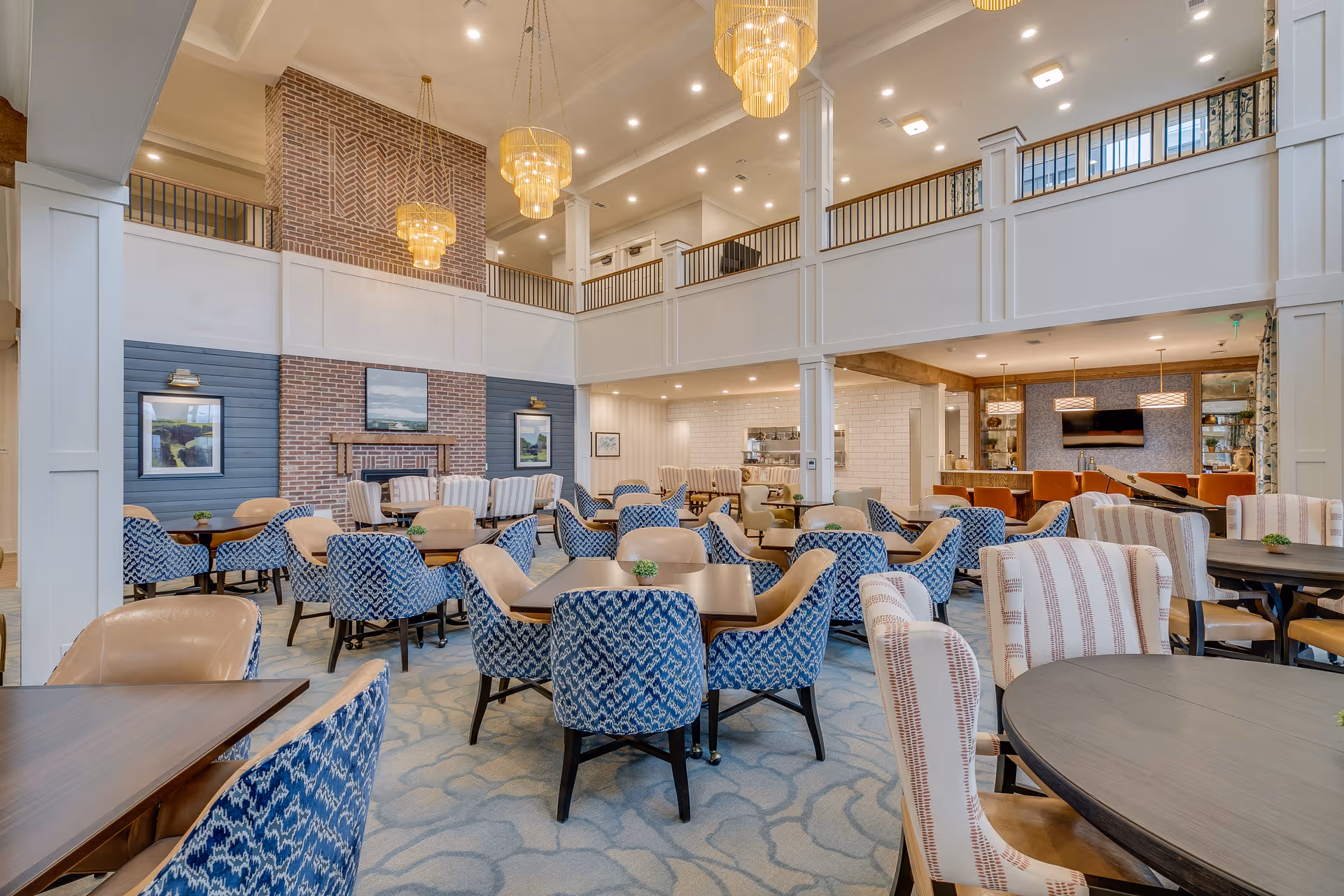 Spacious and elegant dining room with multiple tables and chairs arranged neatly. The room features a high ceiling with large chandeliers, a brick fireplace with framed artwork above it, and a second-floor balcony overlooking the area. The decor includes blue patterned chairs, beige chairs, and small green plants on the tables.