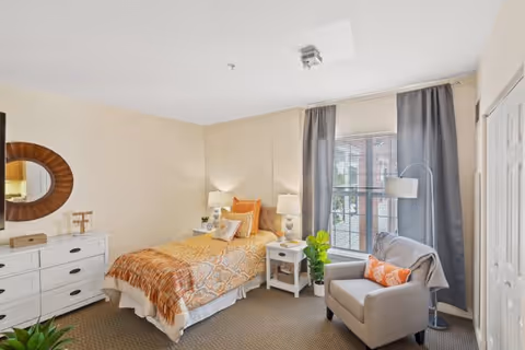 A cozy bedroom with a single bed covered in orange and beige patterned bedding and a brown throw blanket. There is a white nightstand with a lamp and decorative items beside the bed. A comfortable gray armchair with an orange pillow and a gray throw blanket is positioned near a window with gray curtains. A white dresser with a round mirror above it is on the left side of the room. The room has beige walls and carpeted flooring.