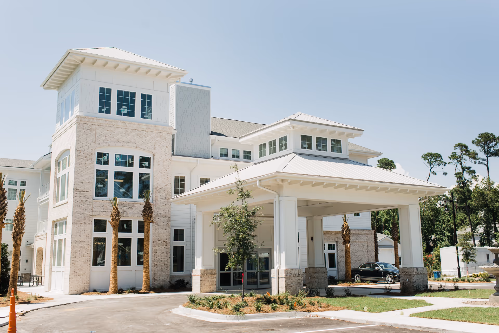 Exterior view of a modern senior living facility building with white brick and siding, large windows, a covered entrance with a peaked roof, palm trees, and a driveway under a clear blue sky.