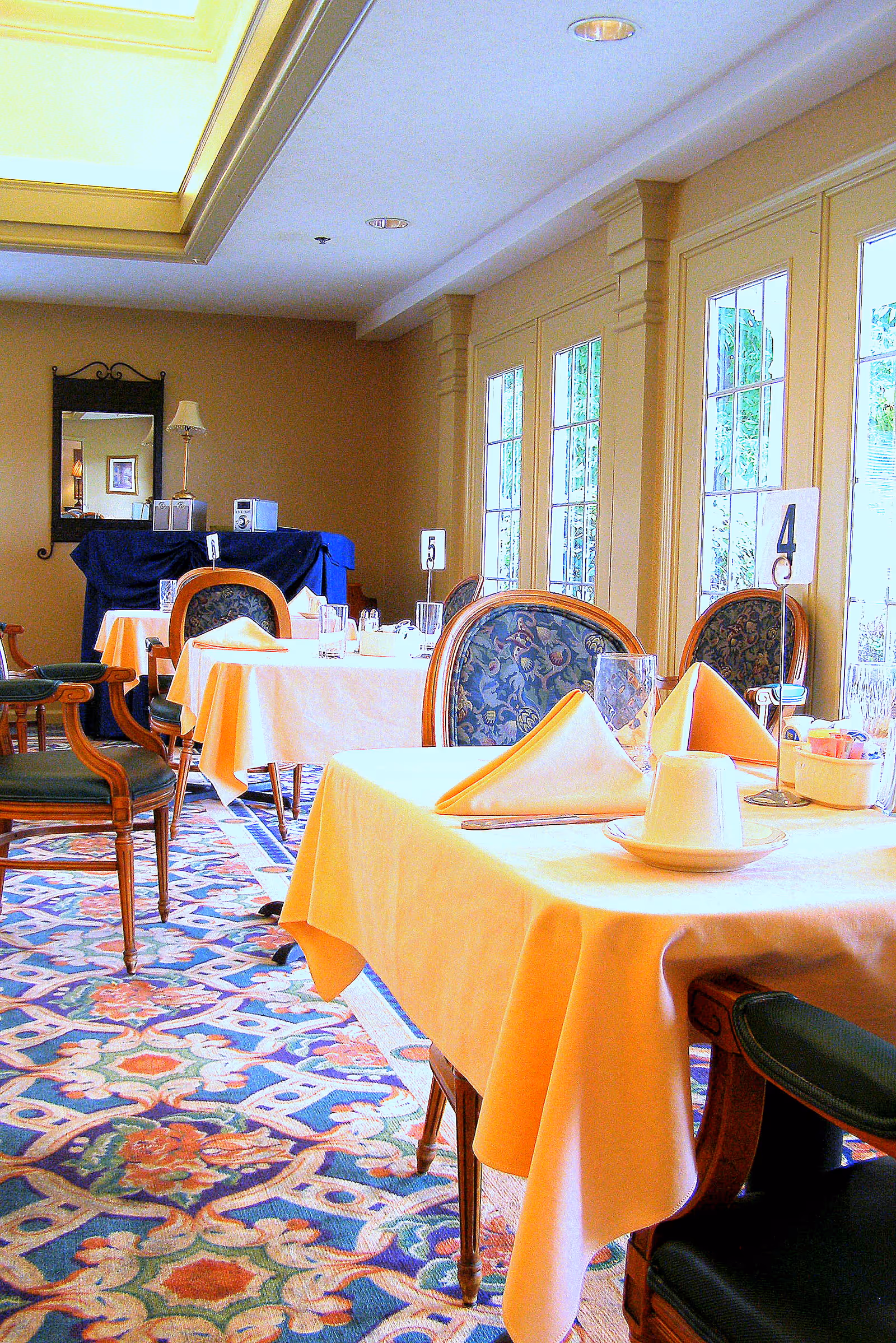 Dining room with tables covered in yellow tablecloths, each set with folded napkins, glasses, and cups. The room has large windows letting in natural light, patterned carpet, and upholstered chairs with floral designs. A mirror and lamp are visible on a table covered with a blue cloth in the background.