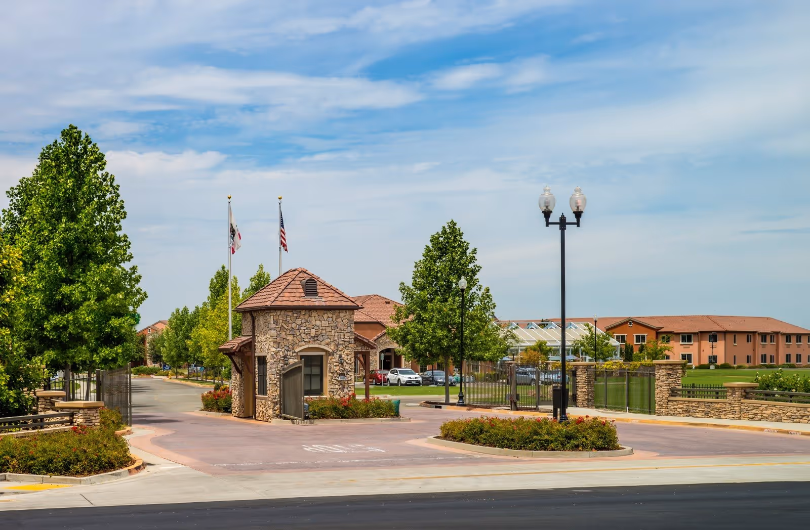 Entrance gate of Eskaton Village Roseville featuring a small stone guardhouse, two flagpoles with flags, surrounding greenery, and a clear blue sky.