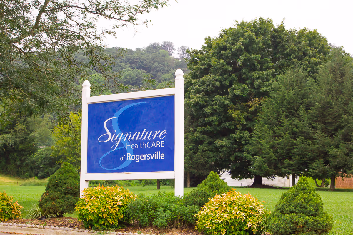 Outdoor view of a blue and white sign for Signature HealthCARE of Rogersville surrounded by green bushes and trees with a grassy area and hills in the background.