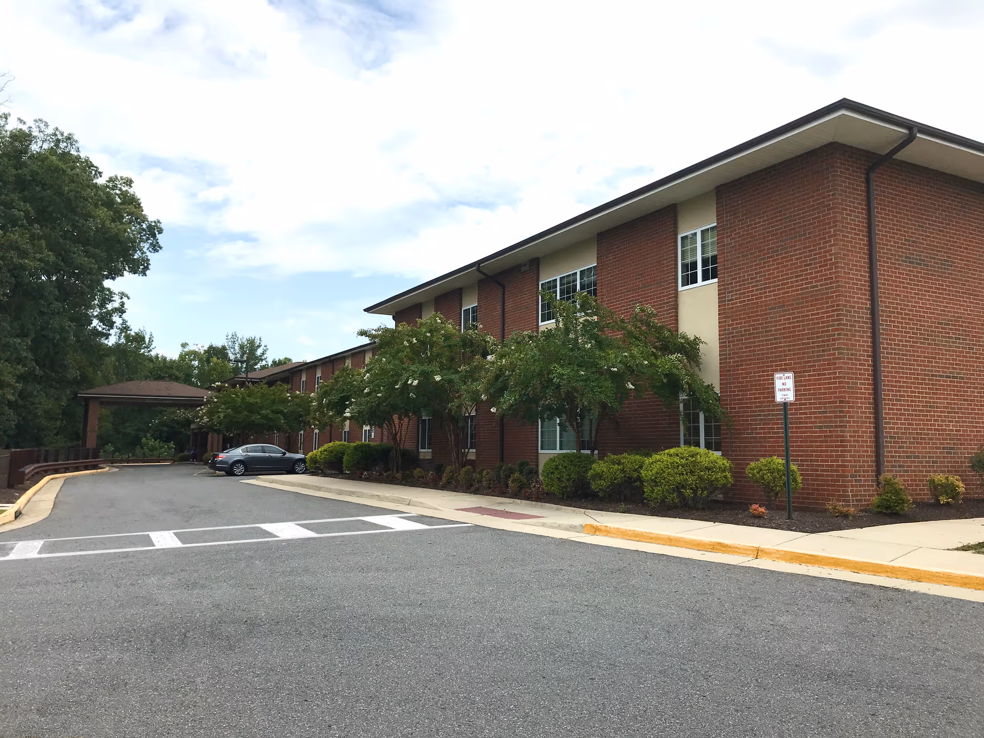 Two-story brick nursing facility with a covered entrance, driveway, parked car, and landscaped shrubs under a cloudy sky.