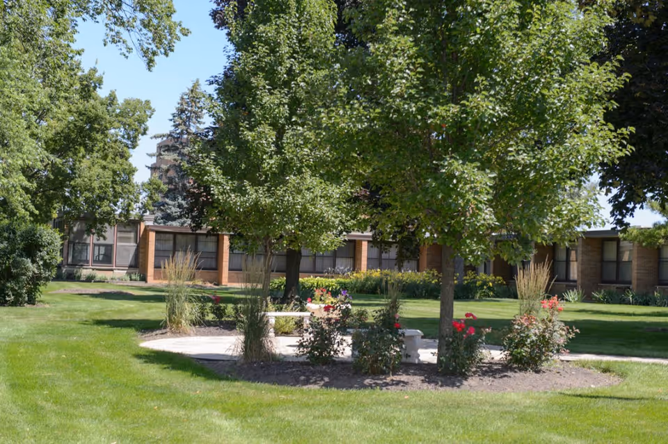A landscaped outdoor area with green grass, trees, and flowering bushes surrounding a circular concrete seating area with benches. In the background, there is a single-story brick building with large windows.
