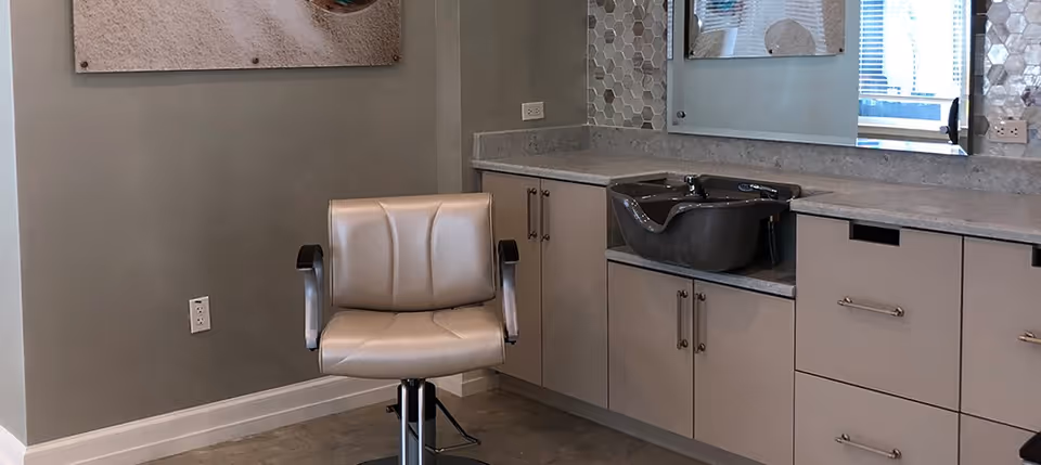 Interior view of a salon area in a senior living facility featuring a beige salon chair in front of a countertop with a built-in black hair washing sink, cabinets below, a large mirror, and a decorative wall with hexagonal tiles.