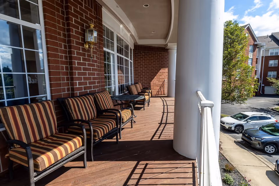 A covered outdoor balcony area with striped cushioned chairs and small tables along a brick wall with large windows. The balcony has large white columns and overlooks a parking lot with several cars and nearby buildings.
