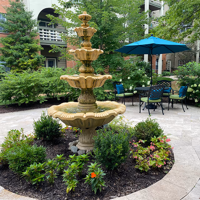 A multi-tiered stone fountain surrounded by landscaped flower beds and patio seating with a blue umbrella in a residential courtyard.