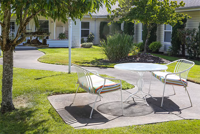 Outdoor patio area with a round white metal table and two white metal chairs with striped cushions, situated on a concrete slab surrounded by green grass and trees, with a building and walkway in the background.