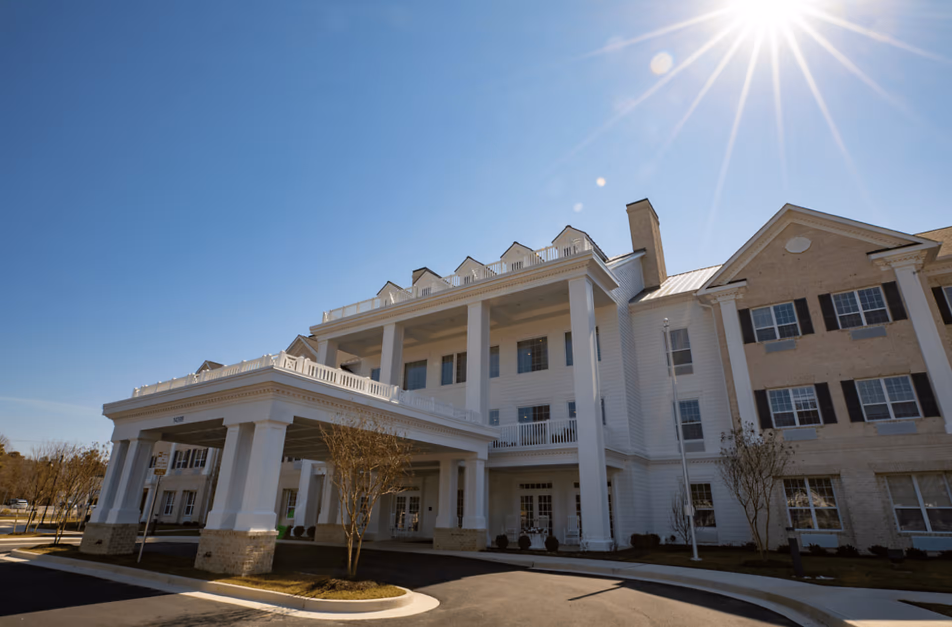 Exterior view of a large senior living facility building with white columns and multiple windows under a clear blue sky with the sun shining brightly.