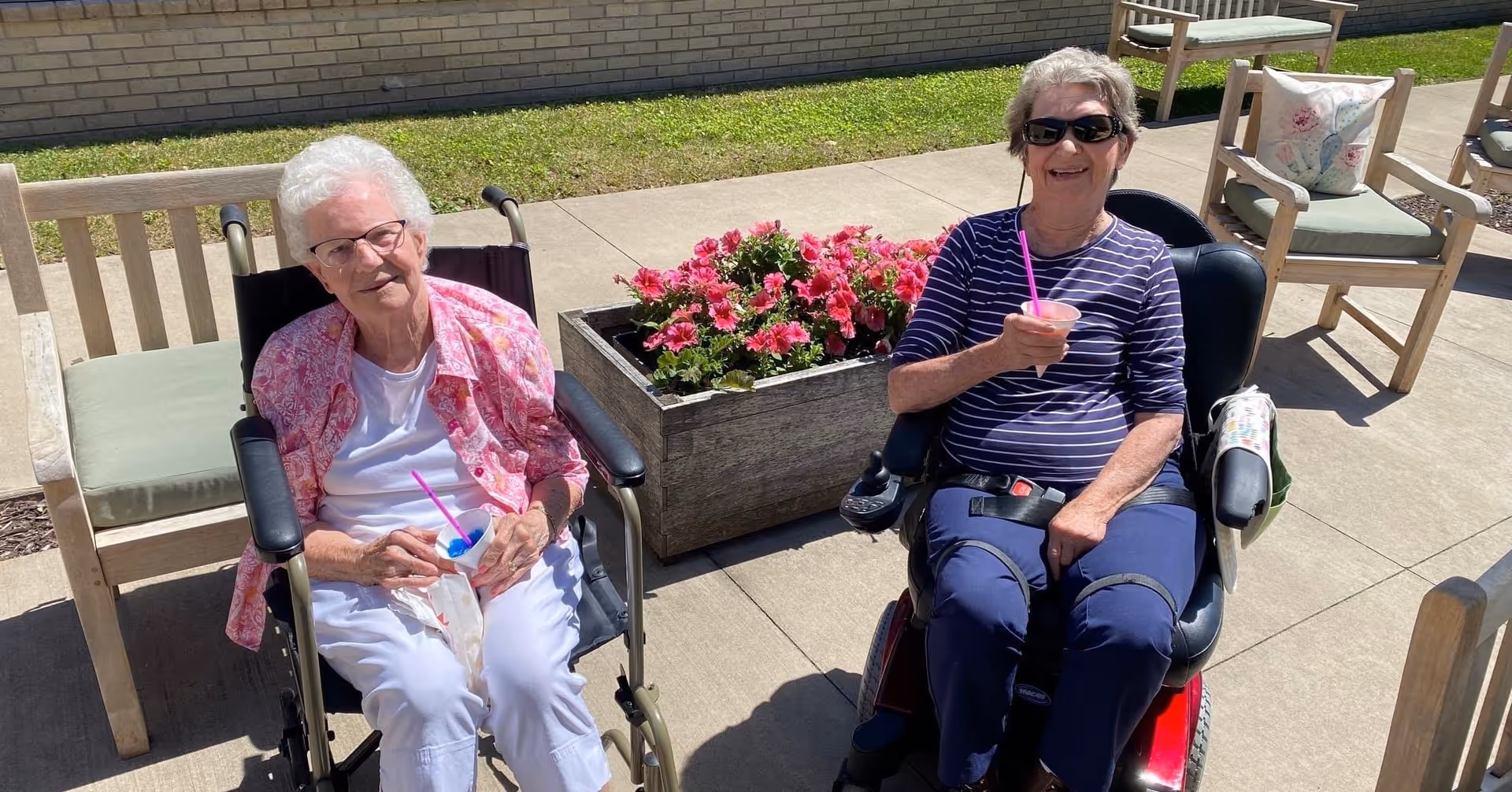 Two elderly women sitting outdoors on a sunny day, one in a wheelchair and the other in a motorized scooter, both holding cups with pink straws. Behind them is a wooden planter box with pink flowers and wooden chairs with cushions on a concrete patio.