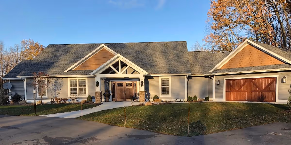 Exterior view of a single-story building with a gray roof and beige siding, featuring a central entrance with double wooden doors and a covered porch. There is a driveway leading up to the entrance and a garage with wooden doors on the right side. Trees with autumn foliage are visible in the background.