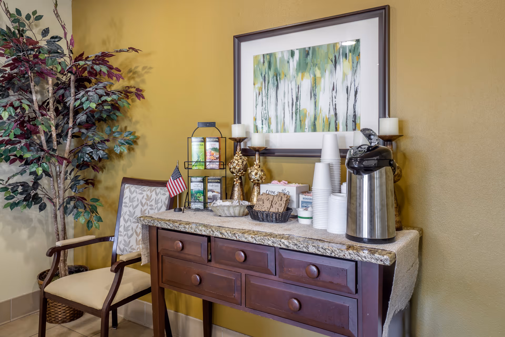 A small refreshment station with a wooden table holding a stainless steel coffee dispenser, stacks of white disposable cups, tea bags in a rack, sugar packets, and decorative candles. A framed abstract painting with green and yellow tones hangs on the mustard yellow wall behind the table. To the left, there is a cushioned wooden chair and a tall artificial plant in a woven basket.