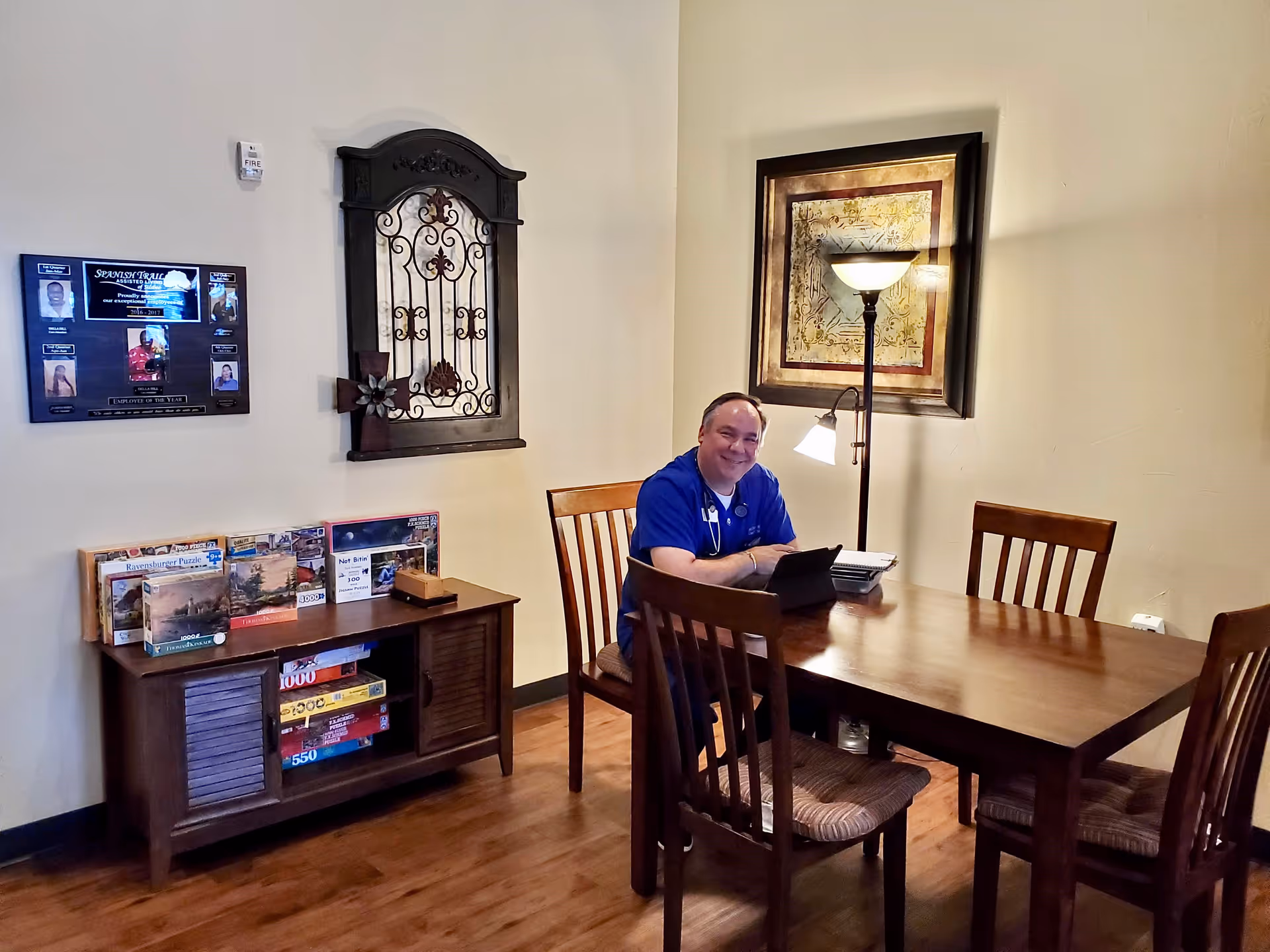 A man in a blue shirt sitting at a wooden table in a room with wooden chairs, a floor lamp, and framed artwork on the wall. There is a wooden cabinet with puzzles and games on it, and a plaque on the wall displaying photos and text related to Spanish Trail Assisted Living of Silsbee.