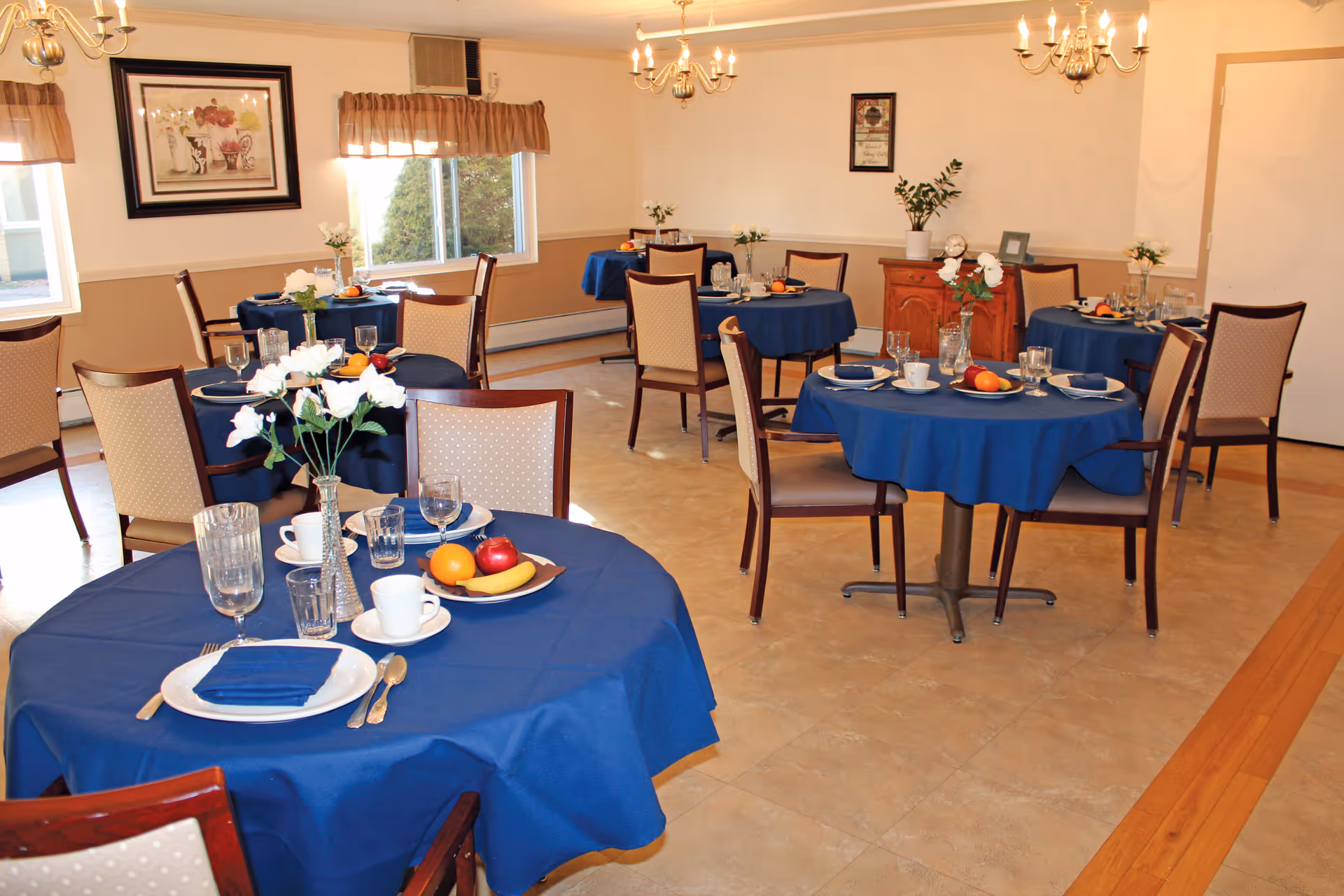 Dining room with multiple round tables covered in blue tablecloths, each set with plates, cups, glasses, silverware, and small flower arrangements. The room has beige walls, windows with brown valances, chandeliers, and framed artwork on the walls.