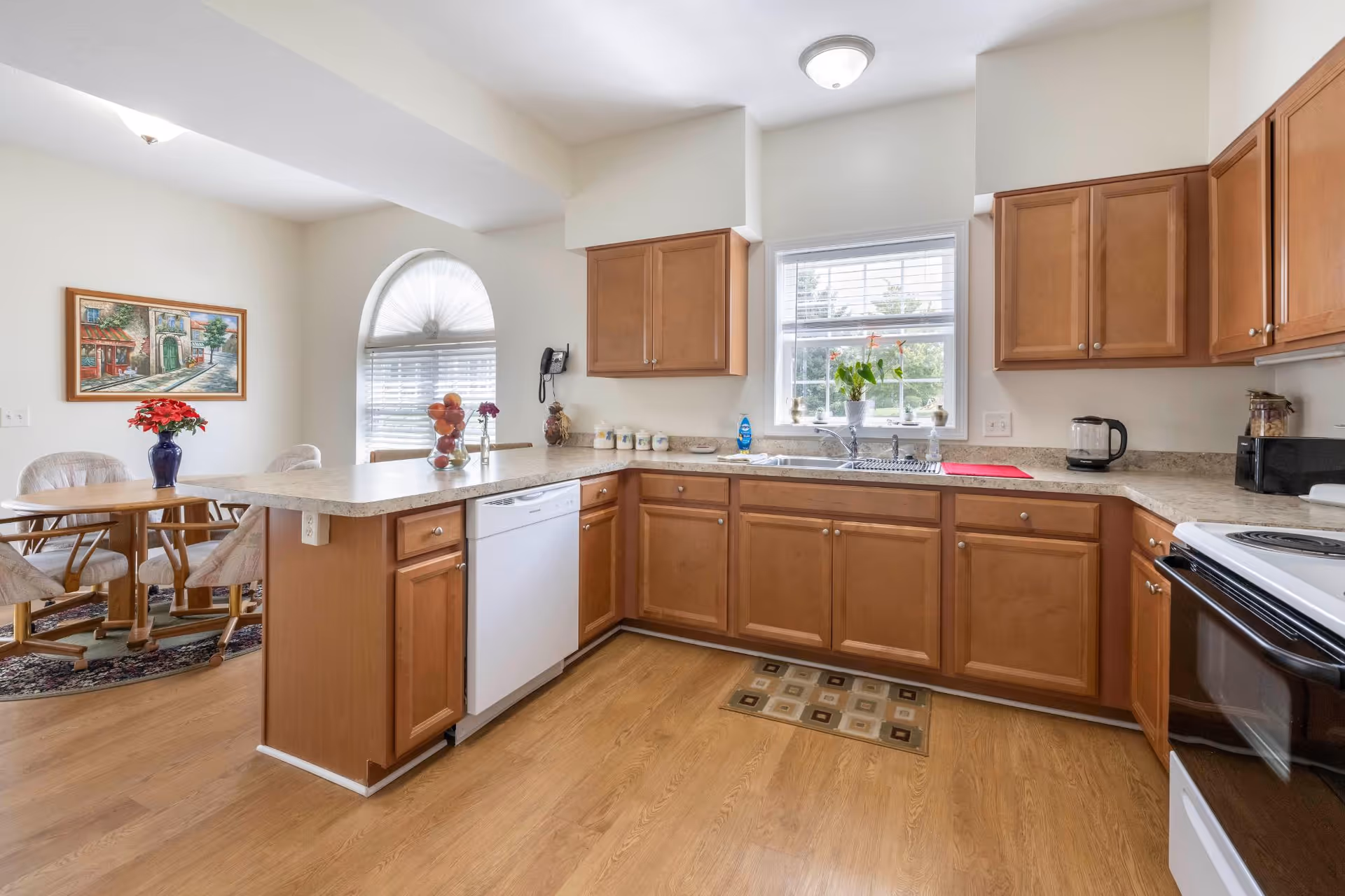 A bright kitchen with wooden cabinets, a white dishwasher, and a white stove. The kitchen has a window above the sink with a plant on the windowsill. There is a countertop with a fruit bowl and a vase with flowers. In the background, there is a dining area with a round wooden table, four chairs, a painting on the wall, and a large arched window with blinds.