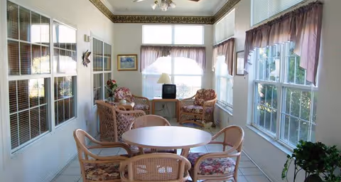 A bright sunroom with large windows on three sides, featuring a round table with four wicker chairs in the foreground and a seating area with two wicker armchairs, a small table, and a TV in the background. The room has tiled floors, floral curtains, and decorative molding near the ceiling.