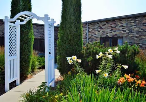 White garden archway over a path among tall evergreens and colorful flowers.