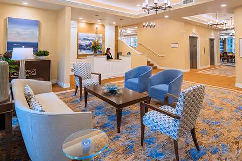 Bright assisted living facility reception lounge with seating area, coffee table, and a staffed front desk.
