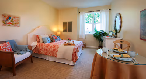 A cozy bedroom in a senior living facility with a bed dressed in red and white patterned bedding, decorative pillows, and a tray with flowers. There is a wooden armchair with cushions on the left, a round table set for two with plates, cups, and napkins on the right, and a window with white curtains letting in natural light. The walls are painted light yellow, and there is a mirror and some framed artwork on the walls.