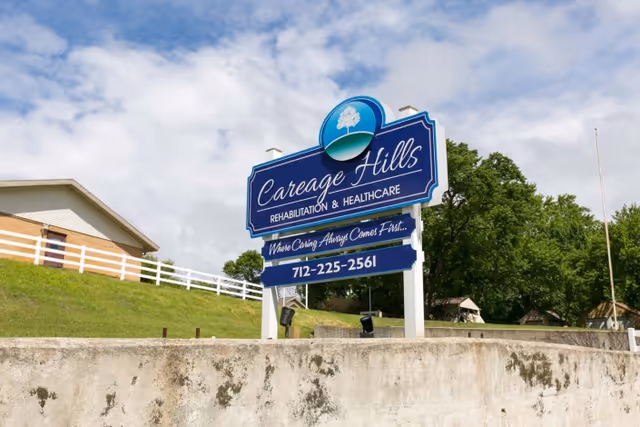 Outdoor view of a large blue and white sign for Careage Hills Rehabilitation & Healthcare with the tagline 'Where Caring Always Comes First' and a phone number, set against a partly cloudy sky and green trees.