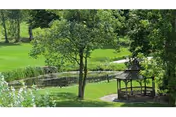 A peaceful outdoor scene featuring a small wooden gazebo surrounded by lush green grass and trees, with a pond and a walking path in the background.