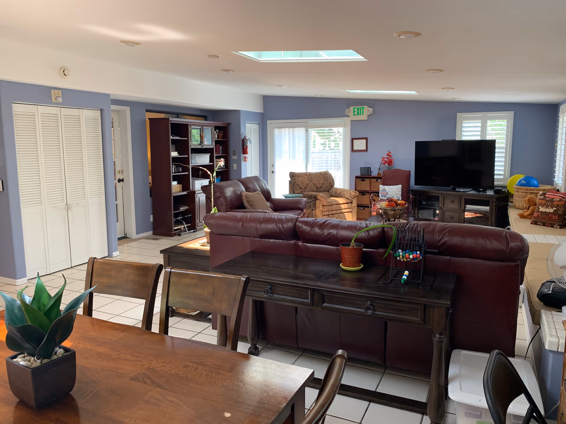 A bright and spacious living room with light blue walls and white tiled floors. The room features a large brown leather sofa, a patterned armchair, a wooden entertainment center with a flat-screen TV, and a wooden dining table with chairs in the foreground. There are skylights on the ceiling, a glass door with an exit sign above it, and various decorative items including plants and cushions.