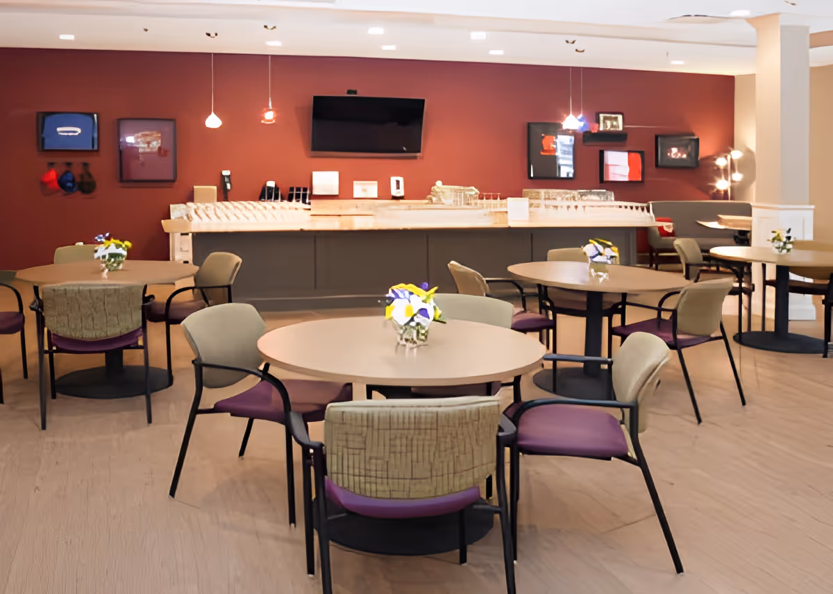 A dining area with round tables and chairs arranged neatly. Each table has a small floral centerpiece. The back wall is painted red and features a mounted flat-screen TV, framed pictures, and pendant lights hanging from the ceiling. There is a counter with various items on it along the back wall.