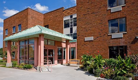 Exterior view of a brick building with a covered entrance supported by pink columns, a bench near the entrance, and landscaped greenery with flowers in front under a blue sky with some clouds.