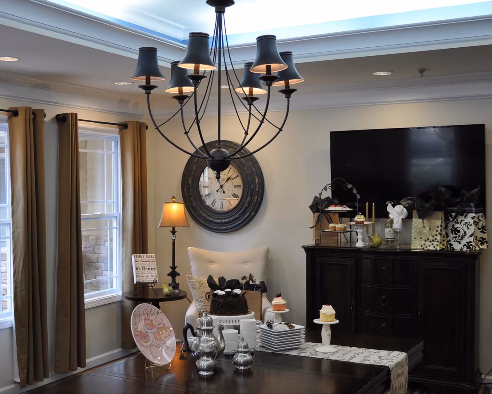 A decorated dining room with a dark wood table set with a silver tea service and cupcakes, a chandelier overhead, and a TV and large clock on the far wall.