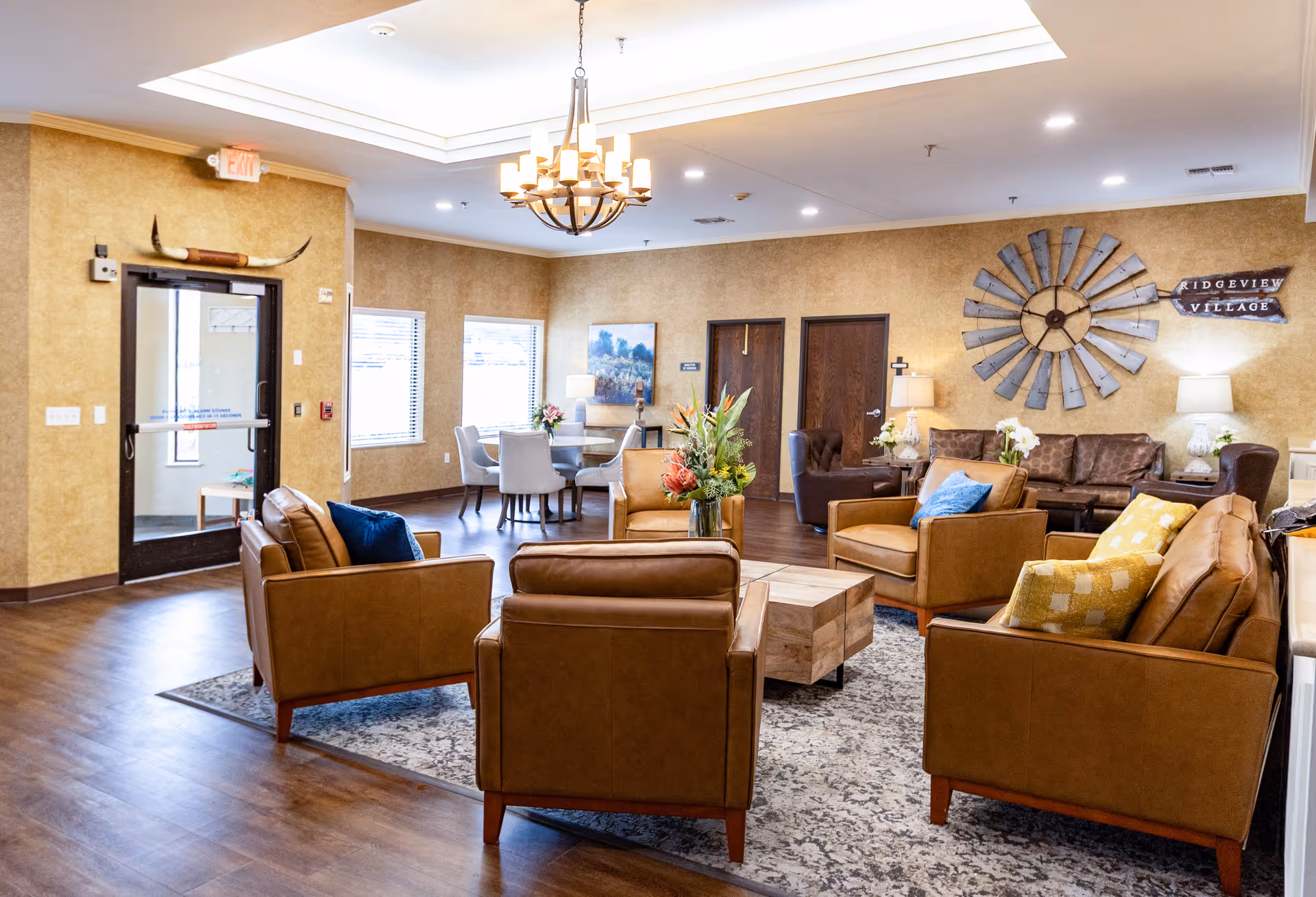 A spacious and well-lit living room area in a senior living facility with multiple brown leather armchairs and sofas arranged around a wooden coffee table on a patterned rug. The walls are beige with decorative elements including a large windmill wall clock and a sign that reads 'Ridgeview Village'. There is a chandelier hanging from the ceiling and a small dining area with a round table and chairs near the windows.