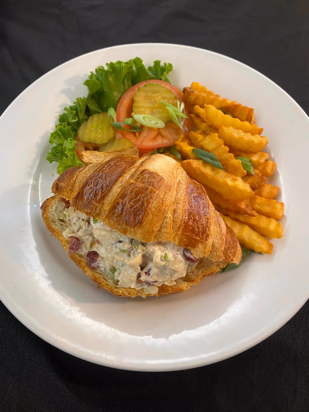 A white plate with a croissant sandwich filled with chicken salad, accompanied by crinkle-cut fries and a side of lettuce, tomato slices, and pickle slices, all placed on a black tablecloth.
