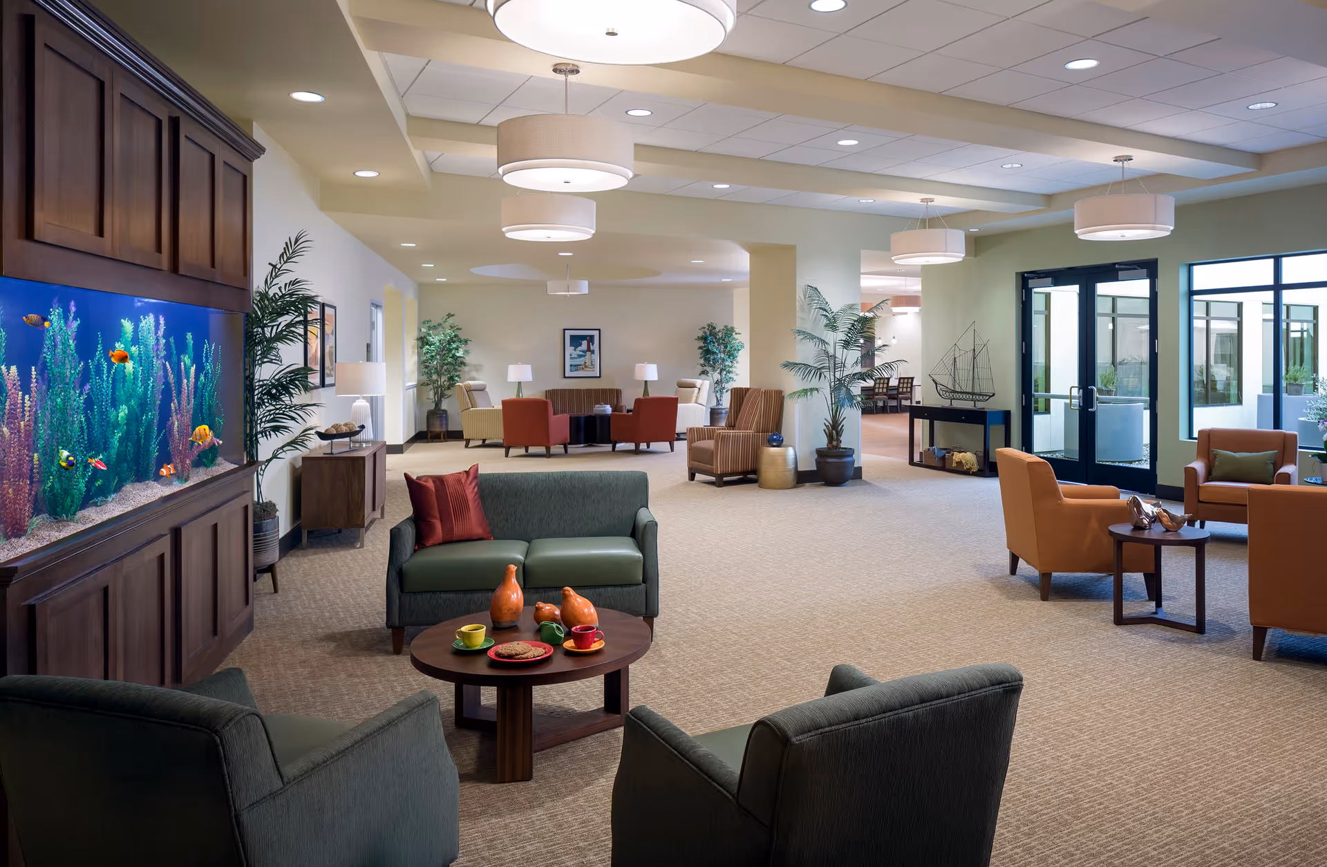 A spacious and well-lit living room area in a senior living facility featuring comfortable seating arrangements including green and orange armchairs and sofas around wooden coffee tables. A large built-in aquarium with colorful fish is on the left wall. The room has beige carpeting, potted plants, modern ceiling lights, and large windows and glass doors letting in natural light.