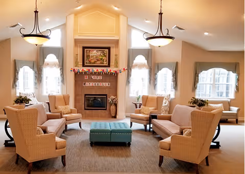 A spacious and well-lit living room with beige walls and carpet. The room features two beige sofas and four beige armchairs arranged around a central teal ottoman. There is a fireplace with a painting above it and two tall windows on either side, each adorned with light blue curtains. Two hanging light fixtures illuminate the room, and there are small plants on side tables near the windows.