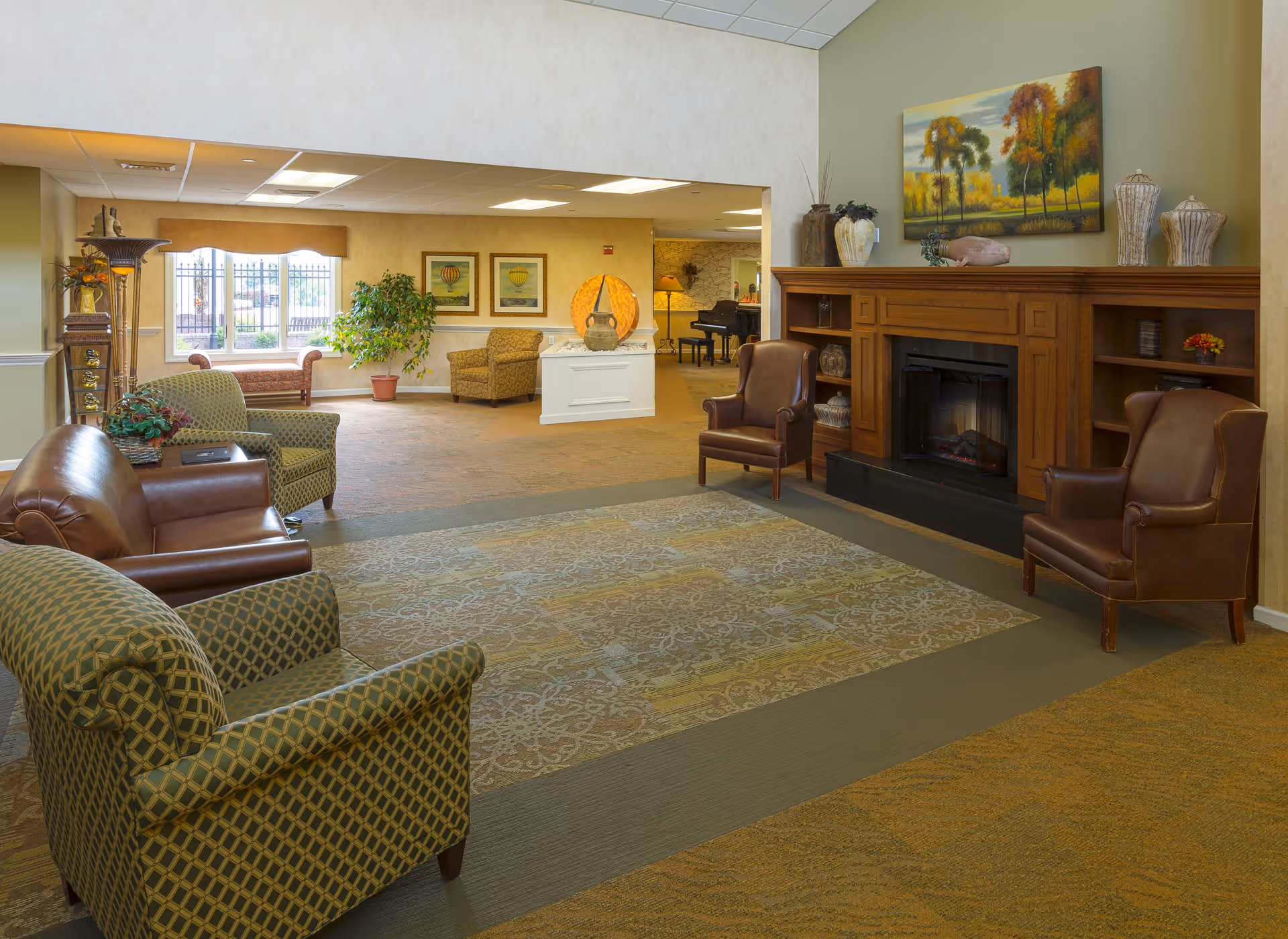 A cozy living room area in a senior living facility with a wooden fireplace, two brown leather armchairs, two patterned armchairs, a leather sofa, and a large decorative rug. The room is decorated with vases and a painting of autumn trees above the fireplace. In the background, there is a window with a bench, framed artwork on the walls, a potted plant, and a piano in an adjacent room.