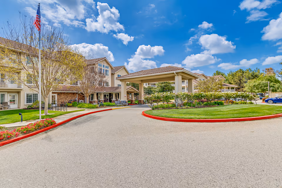 Front entrance of a senior living building with a covered porte-cochère, landscaped circular driveway, and an American flag under a blue sky.