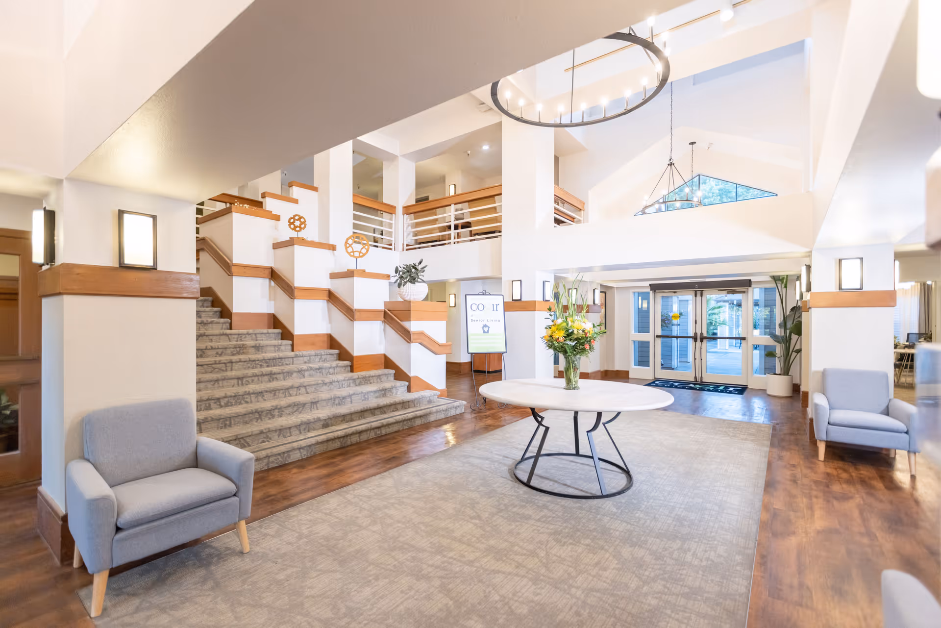 Bright and spacious lobby area with a large staircase, two gray armchairs, a round table with a flower arrangement in the center, and large windows near the entrance letting in natural light.