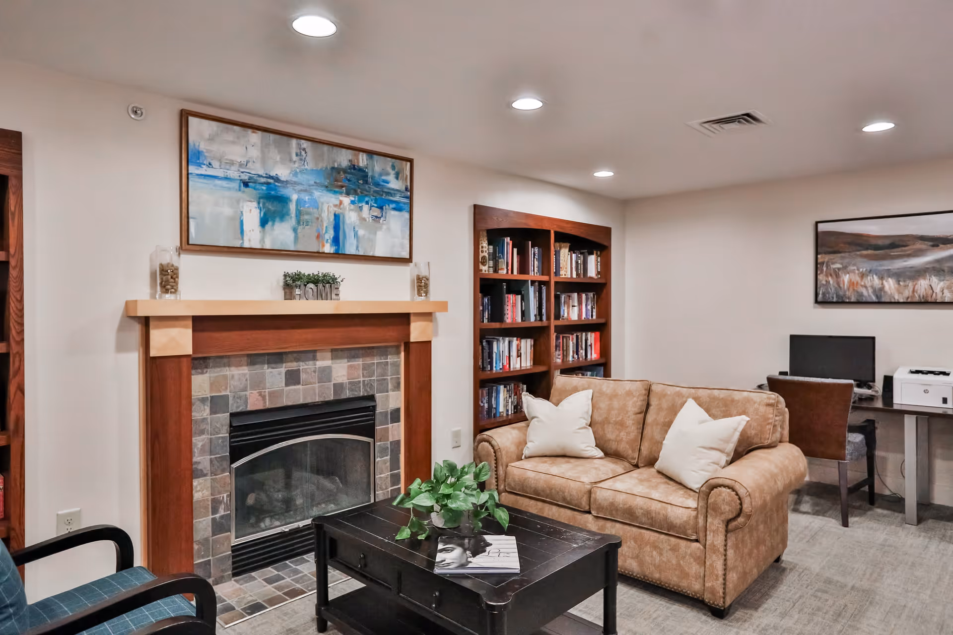A cozy living room area with a tan leather sofa adorned with two white pillows, a black coffee table with a plant and a magazine, a wooden bookshelf filled with books, a fireplace with a tiled surround and a mantle decorated with a small plant and two glass jars, and a desk with a computer and printer in the background. The walls are white with two framed paintings, and the ceiling has recessed lighting.