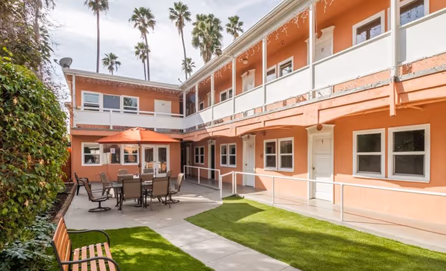 Outdoor courtyard area of a senior living facility with peach-colored two-story building walls, several windows and doors, a patio table with chairs under an orange umbrella, a wooden bench, green artificial grass, and palm trees in the background under a partly cloudy sky.