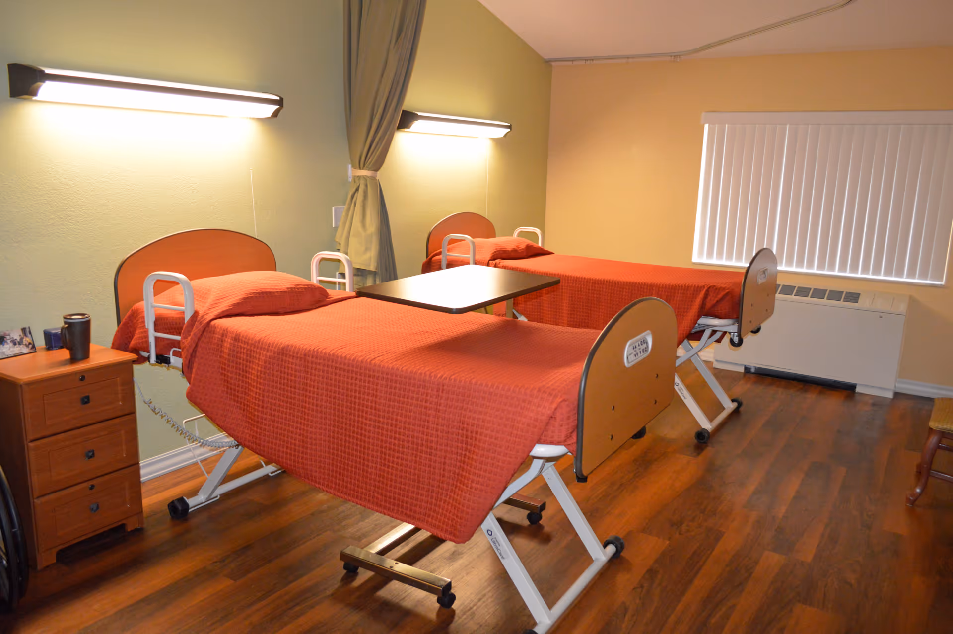 A room with two hospital-style beds covered with red blankets, each with a headboard and side rails. There is a small wooden nightstand with drawers and a cup on it next to one bed. A curtain is tied back between the beds, and there are wall-mounted lights above each bed. The room has wooden flooring and a window with vertical blinds.