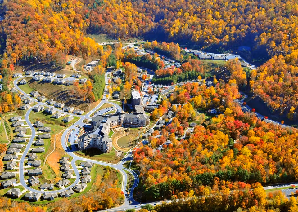 Aerial view of Givens Estates senior living community surrounded by dense forest with autumn foliage. The image shows multiple residential buildings, winding roads, parking areas, and landscaped grounds nestled in a valley with colorful fall trees.