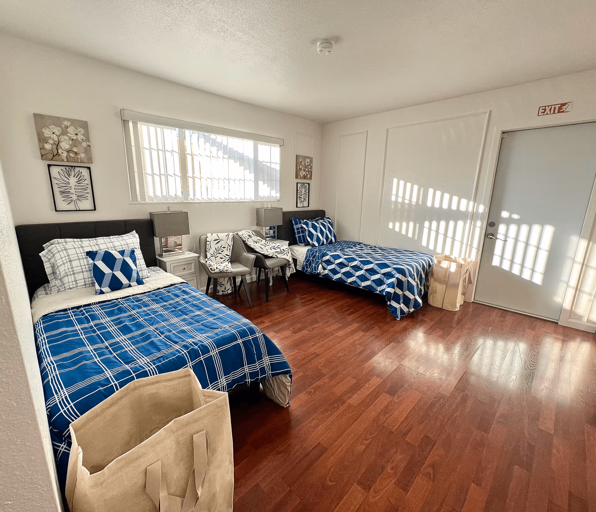 Bright shared bedroom with two twin beds with blue patterned bedding, a nightstand and chairs on hardwood floors, sunlight streaming through a window and an exit door.